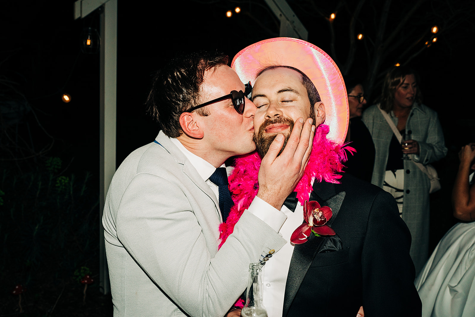groom in a pink cowboy had and pink boa being kissed on the cheek by a friend